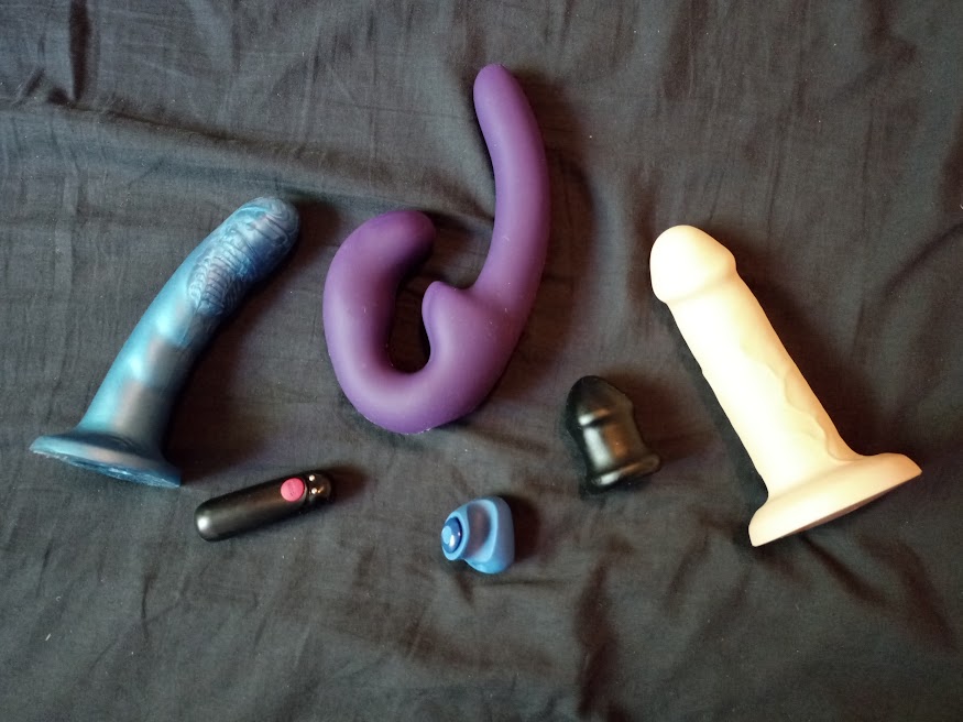 2 flared base dildos, one feeldoe, one bullet vibe, transcock stroker, and finger vibe sit laid out on wrinkled black sheet.